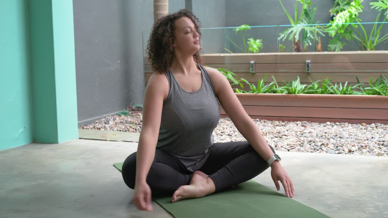 Mulher afro-americana seguindo a instrução, alongando-se e respirando em um colchonete de ioga verde em um pátio coberto