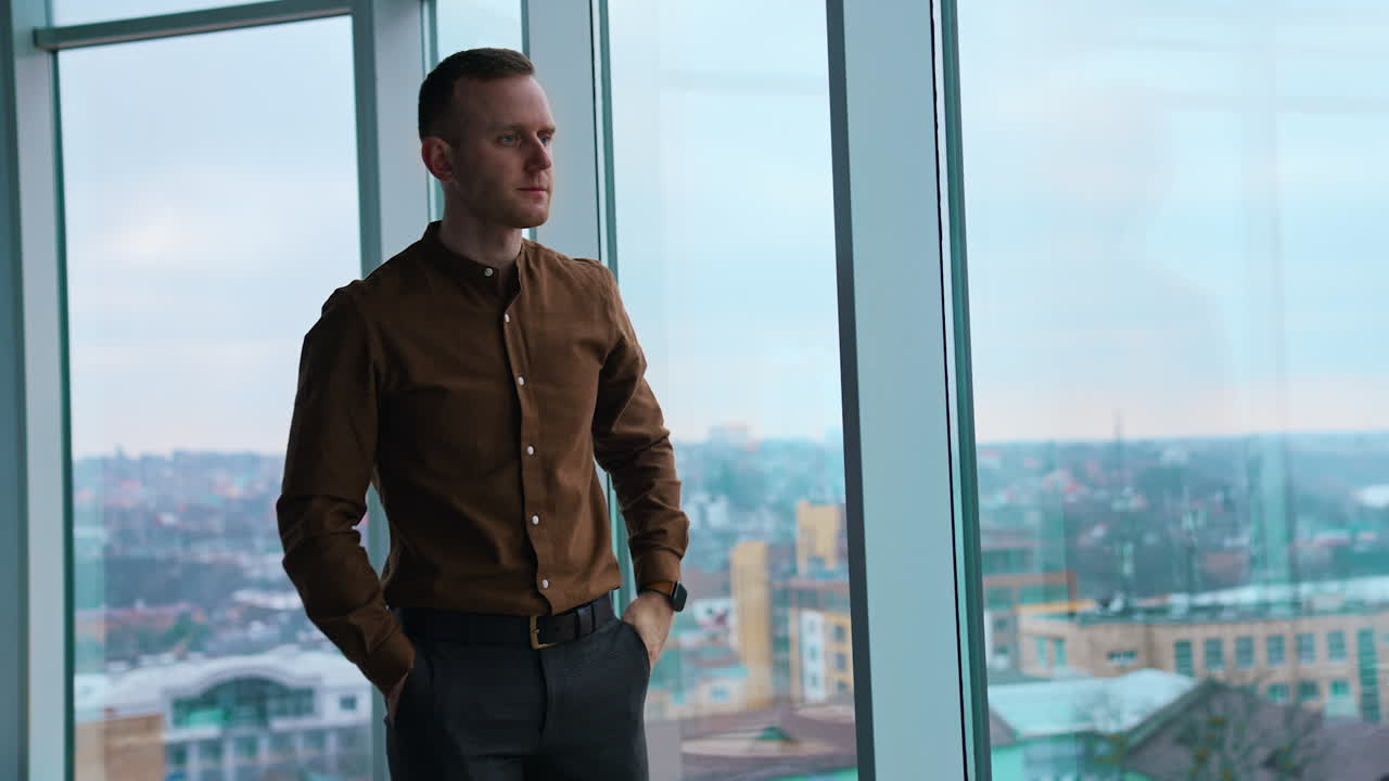 Full-length portrait of a businessman indoors. Tired young man walking along the office and stands by the large window with city view background.