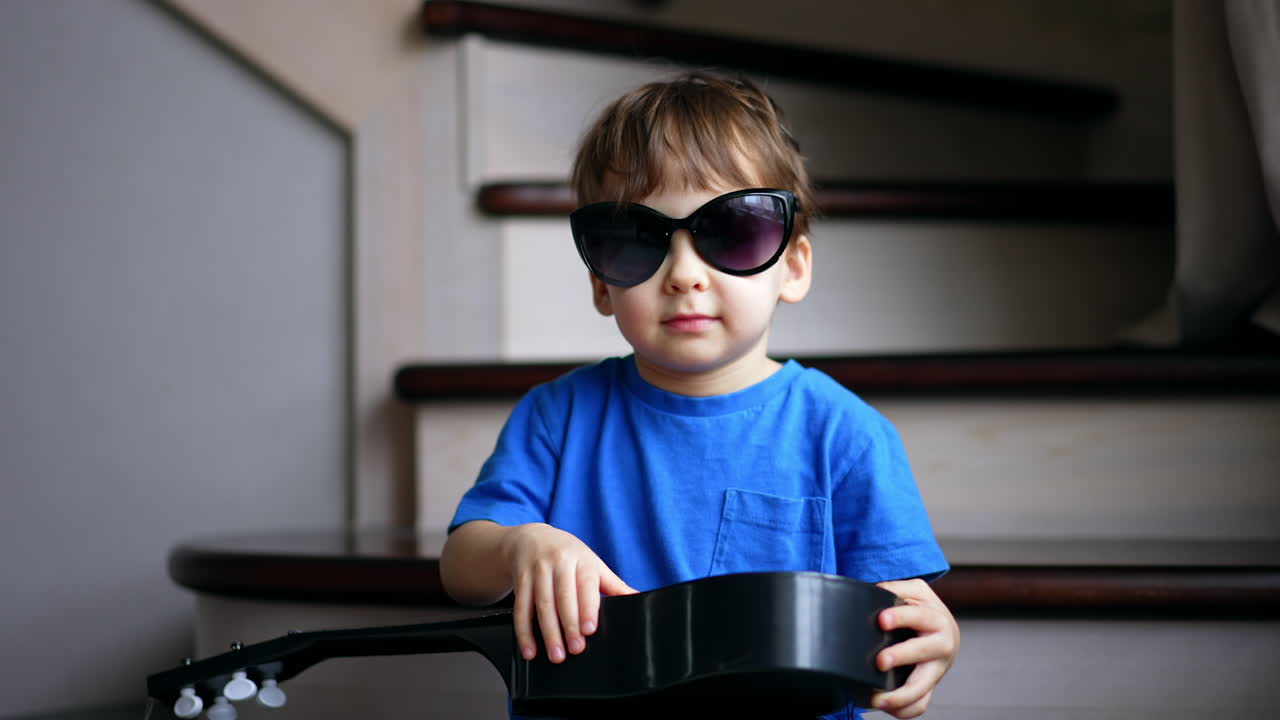 Beautiful baby boy wearing mom's sunglasses sits holding a guitar. Toddler having fun at home.