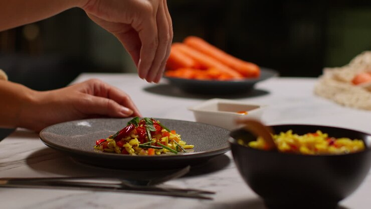 Gros plan sur une femme chez elle dans la cuisine préparant un repas végétarien ou végétalien sain, saupoudrant des herbes sur des pâtes orzo et des tomates rôties
