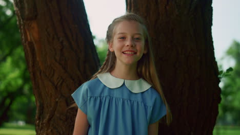 joyful little girl pose at tree trunk. smiling kid with braid look in camera.