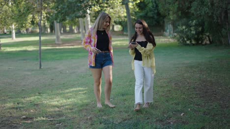 two women walking in park using smartphones