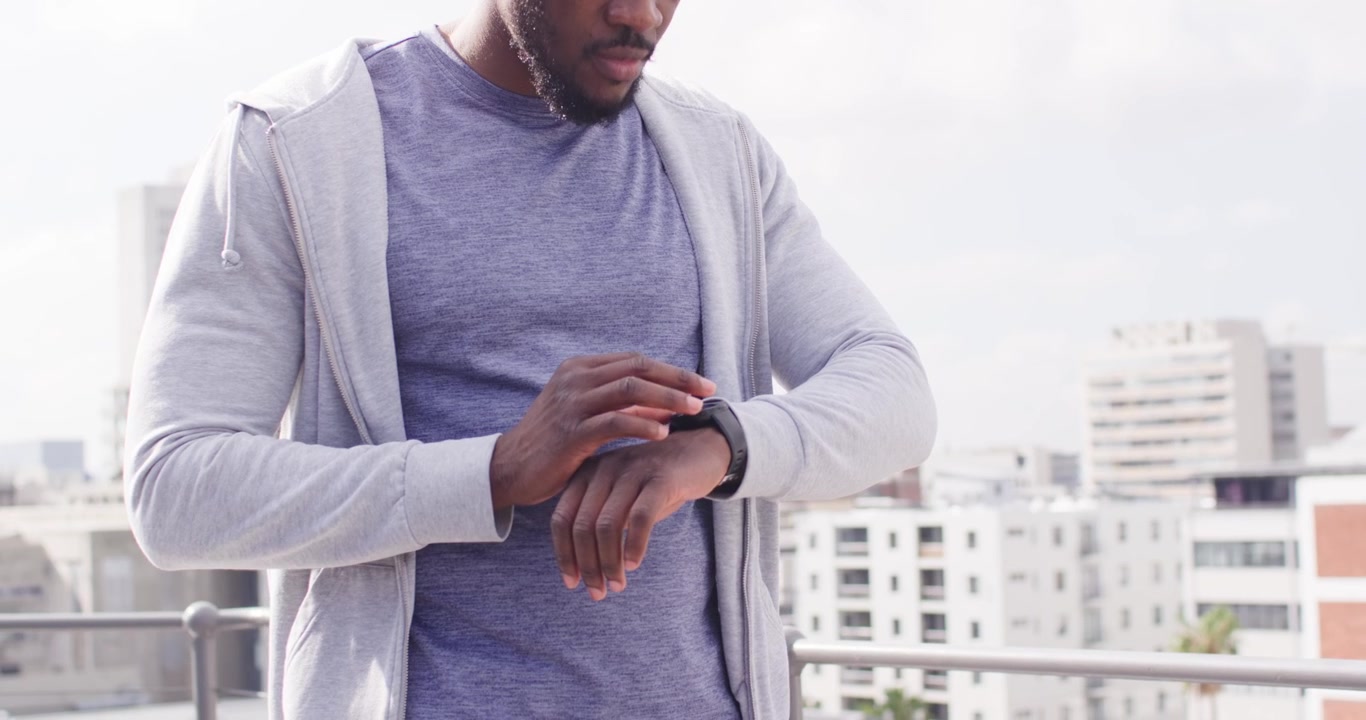 Adult African man warming up on rooftop after checking black smartwatch in gray hoodie, dark shorts