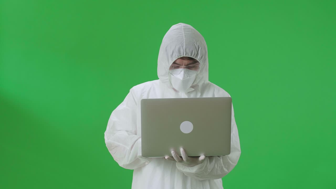 Asian Man Wearing Personal Protective Equipment Uniform Ppe, Medical Face Mask, Holding And Working With Laptop Computer In The Green Screen Studio