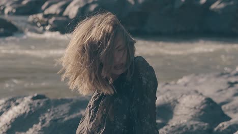 woman in dress sits on large stone near river on windy day