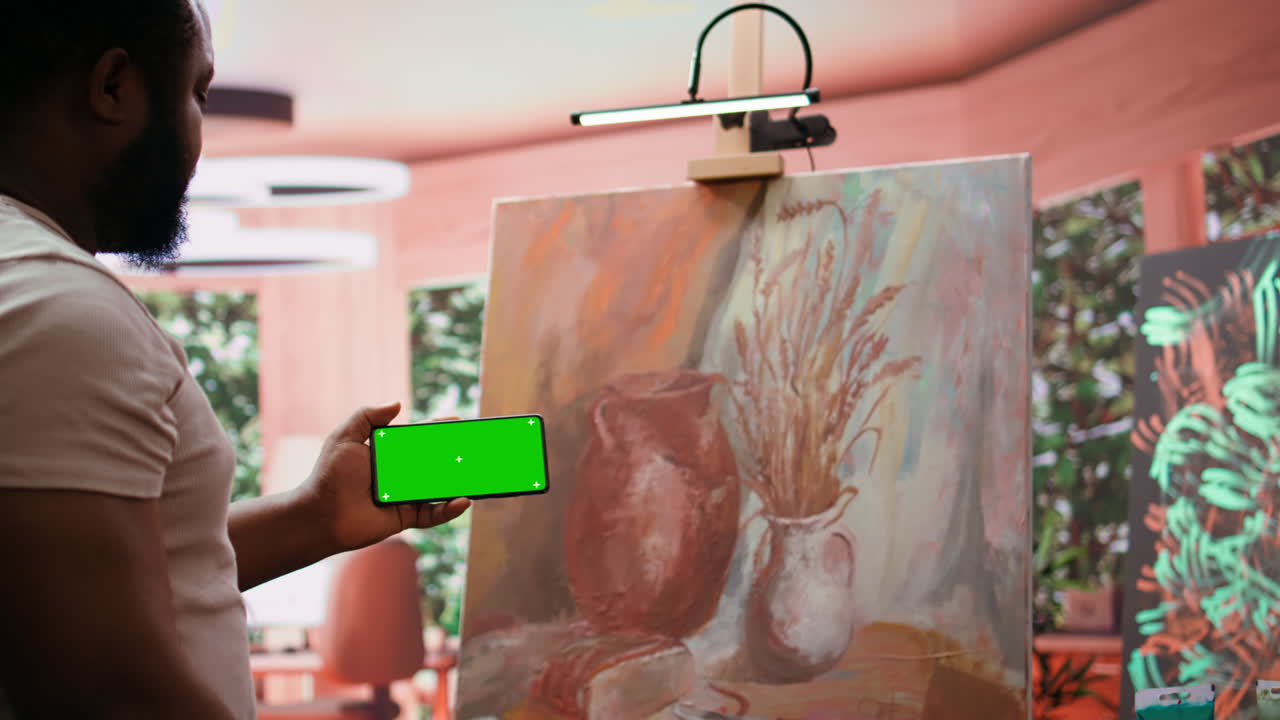 Man holding a phone with a green screen next to a still life painting