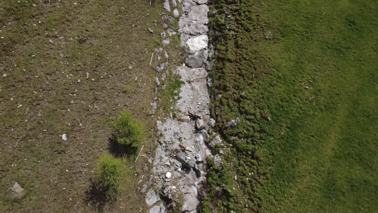 A narrow alpine stream flows through a green mountain valley with scattered rocks and trees.