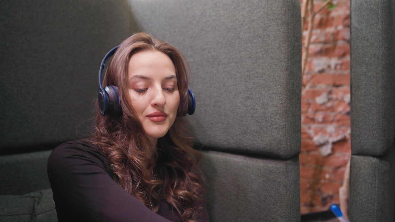 Light skin lady with blond hair gently nodding her head with headset on, seated in a quiet, cushioned lounge corner. She appears relaxed and immersed in music with eyes softly closed