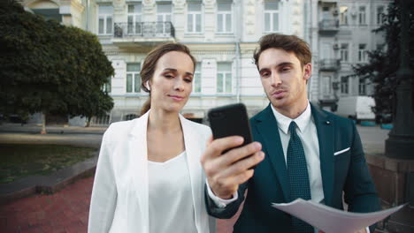 Friendly-business-man-showing-smiling-woman-information-on-phone-on-city-walk.