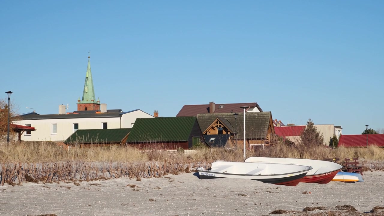 Traditionelle hölzerne Fischerboote liegen am Sandstrand von Kuźnica, Halbinsel Hel, mit Küstenhäusern an der Ostsee im Hintergrund unter einem ruhigen Morgenhimmel.