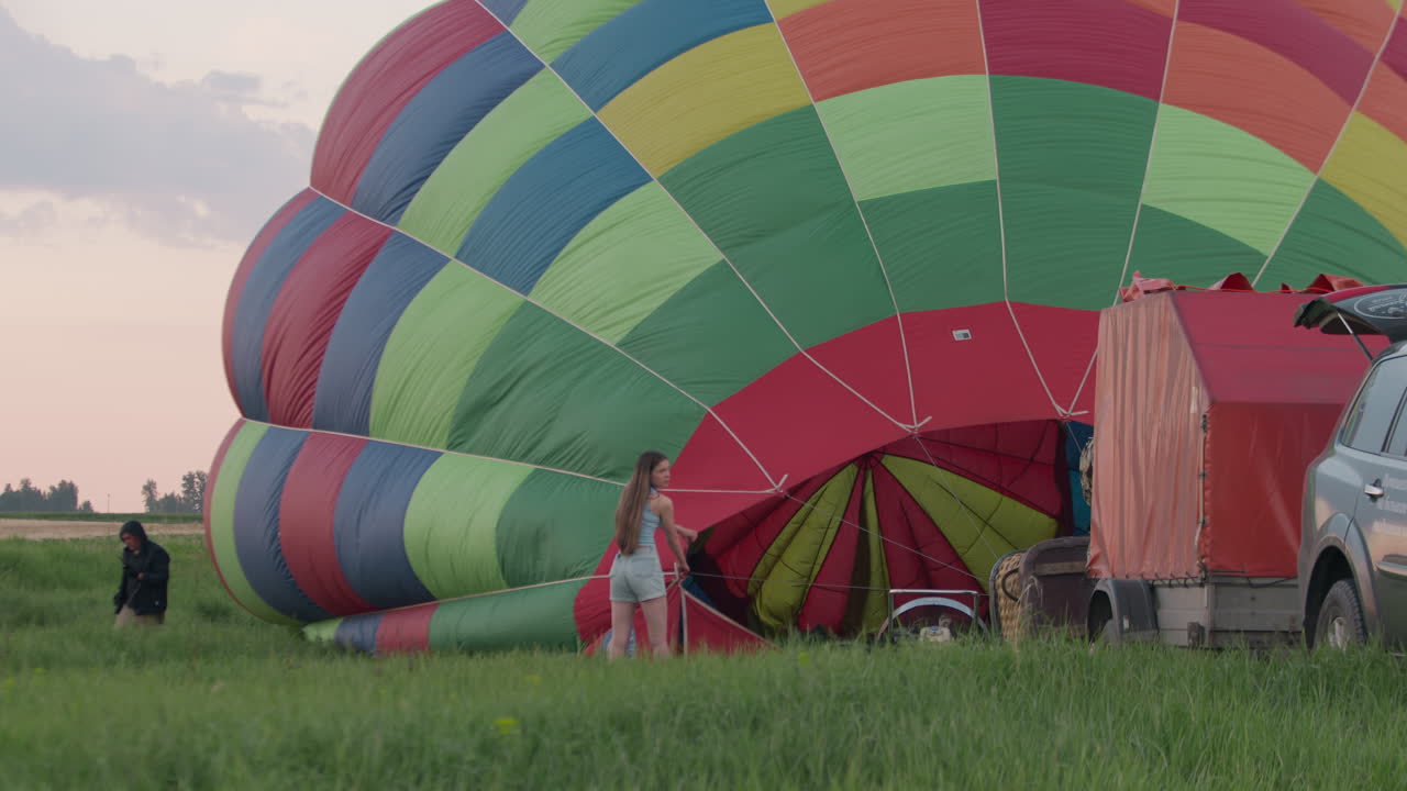 Woman stands near colorful hot air balloon envelope, gripping rope during early stage of inflation while another person approaches from behind near vehicle and equipment trailer on green open field