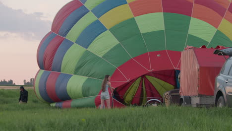 woman stands near colorful hot air balloon envelope, gripping rope during early stage of inflation while another person approaches from behind near vehicle and equipment trailer on green open field