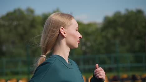 close up of lady with hair tied back in green sweater jogging in outdoor volleyball court with volleyball net and trees in background, capturing athletic movement in fitness training