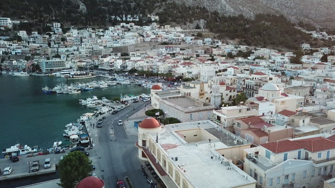Greece,Kalimnos, Aerial footage of the city center, starting from the harbor, goes along the seaside road, flies towards to the clock tower and the church behind the iconic municipality building,