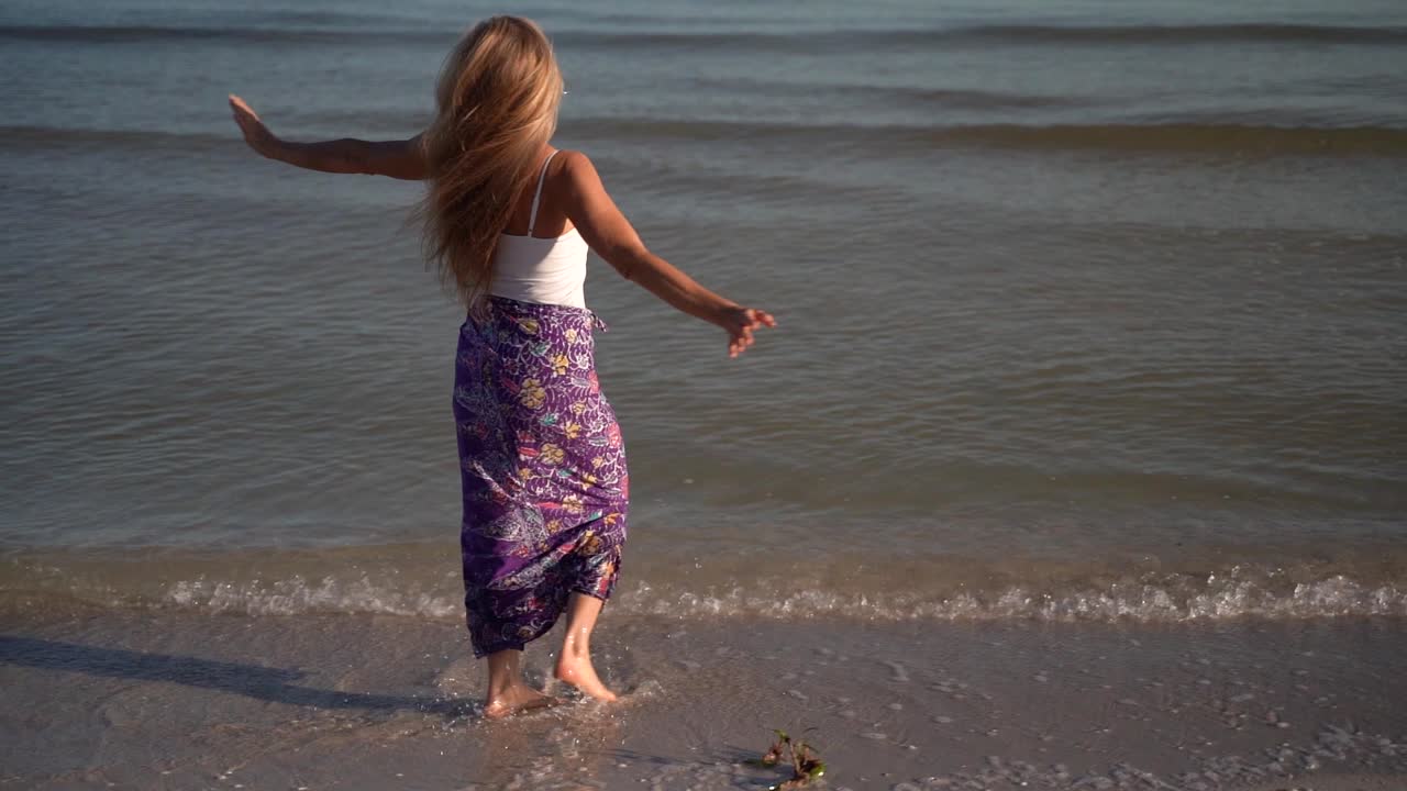 자유를 축하하는 팔을 뻗은 채 물에 튀는 해변에서 사롱을 입은 성숙한 여인의 근접 촬영 슬로우 모션