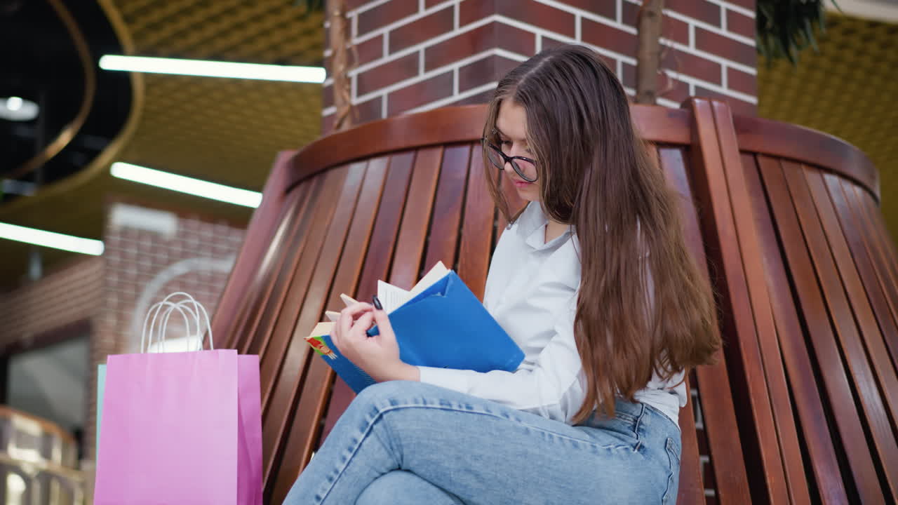 mujer sentada ligeramente doblada, piernas cruzadas, hojeando un libro, con una bolsa de compras cerca en un entorno de centro comercial moderno, mostrando una actividad interior atractiva