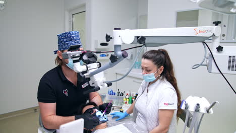 male doctor in dark uniform looks at endodontic microscope performing dental procedure. female nurse sitting beside watching the process.