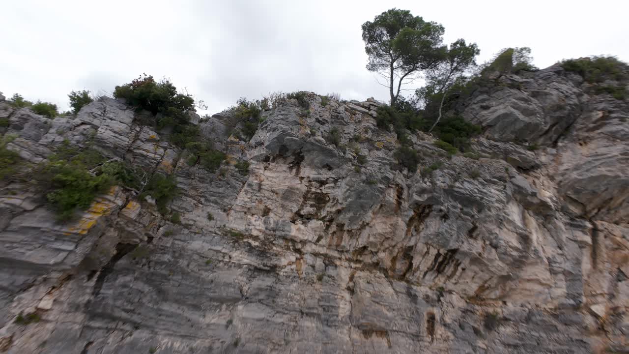 Dynamic FPV drone footage flies along rugged limestone cliffs covered in green vegetation, Ligurian Sea, Noli, Italy. Aerial