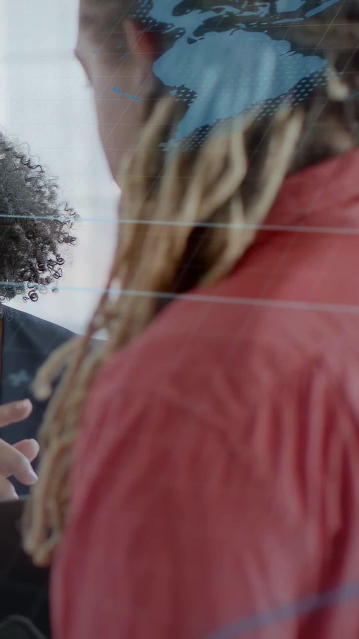 Video verticale: Uomo con giacca rossa che ascolta mentre una donna entra, indicando una mappa su vetro durante una conversazione in ufficio.