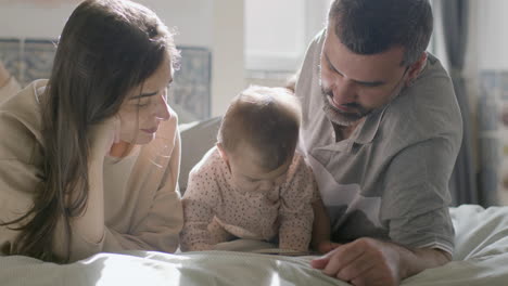 padres amorosos y su linda niña acostada en la cama y viendo dibujos animados en una tableta digital el domingo por la mañana