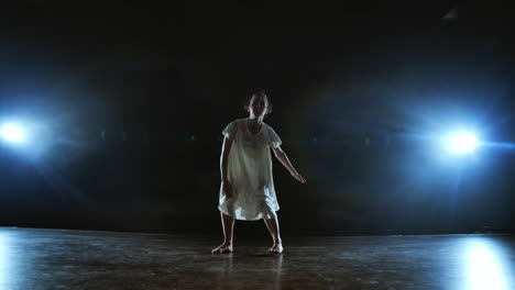 a young girl in a white dress dances and makes rotations in slow motion on the stage with smoke in spotlights in a full shot.