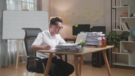 hombre asiático mirando la computadora portátil y tomando notas mientras trabaja con documentos en la oficina