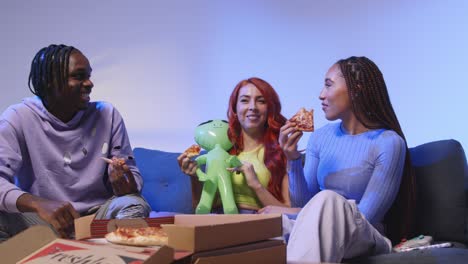 group of gen z friends sitting on sofa at home eating takeaway pizza and playing with toy alien