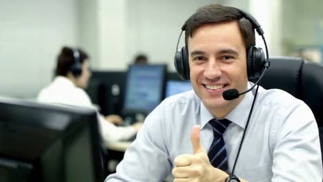 a man wearing a headset and smiling at the camera in call center, thumbs up.