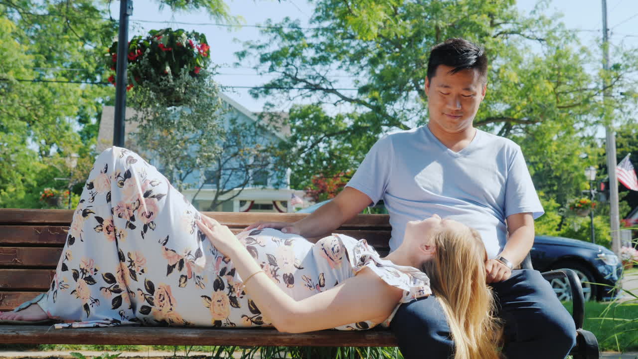 임신한 아내를 둔 아시아 남자가 함께 공원 벤치에서 쉬고 있습니다.