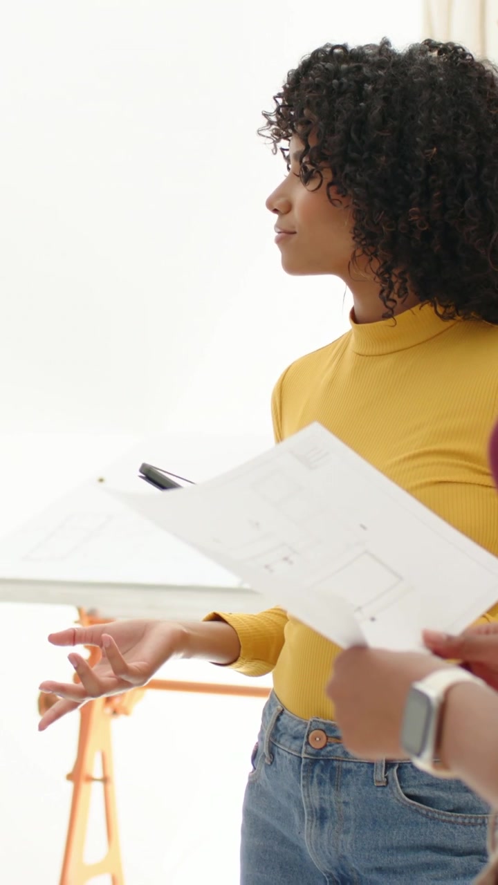Vertical video: Pullback shot showing woman in maroon reviewing plans in studio with peer pointing