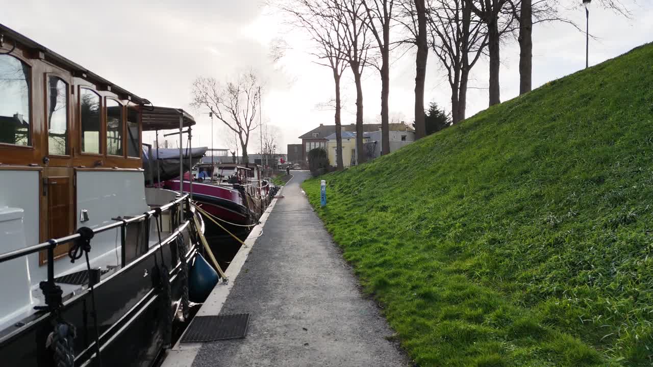 Low aerial tracking along a path with boats on one side and embankment on the other