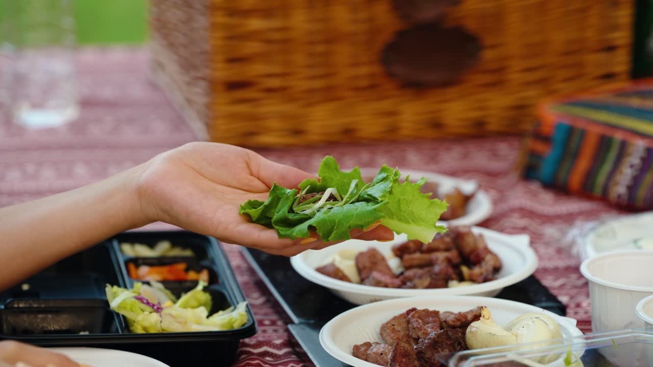A woman's hands prepare a traditional Korean barbecue lettuce wrap (ssam) by adding grilled pork, garlic, and sauce to a fresh lettuce leaf during an outdoor picnic with friends or family