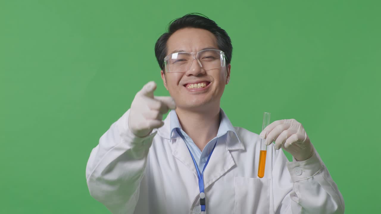 Close Up Of Asian Man Scientist With Orange Liquid In The Test Tube Smiling And Touching His Chest Then Pointing At You While Standing On The Green Screen Background In The Laboratory