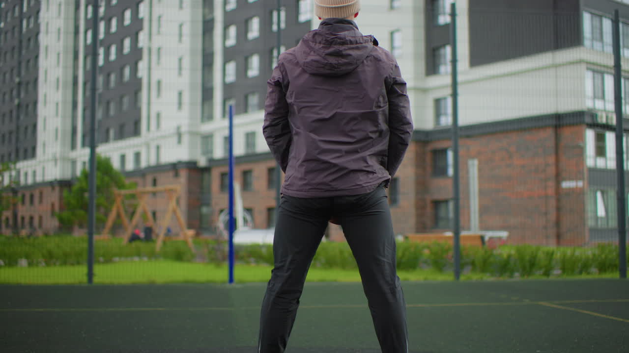 Mand laver squats udendørs nær lejligheder iført vinterjakke og hue, og udfører langsomme, kontrollerede gentagelser på en legeplads med gummibelægning foran en moderne lejlighedsbygning i trænerstil