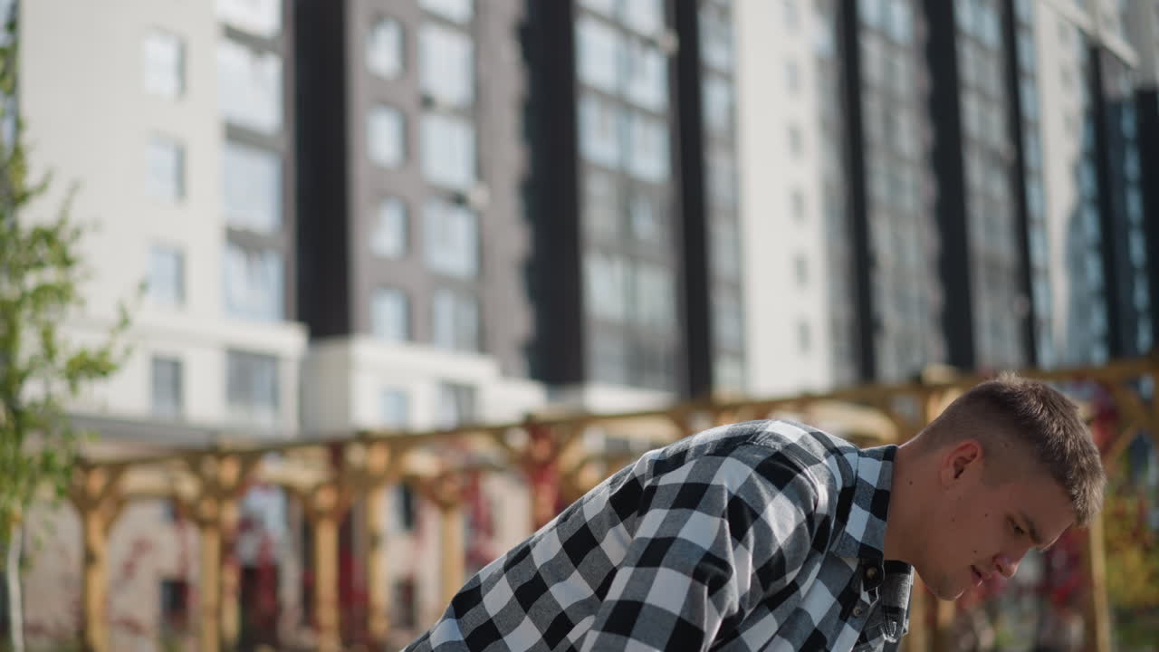 calm fair man in checkered shirt sits under bright sun and takes pill from front pocket surrounded by modern high-rise buildings wooden pergola and green plants on peaceful city day
