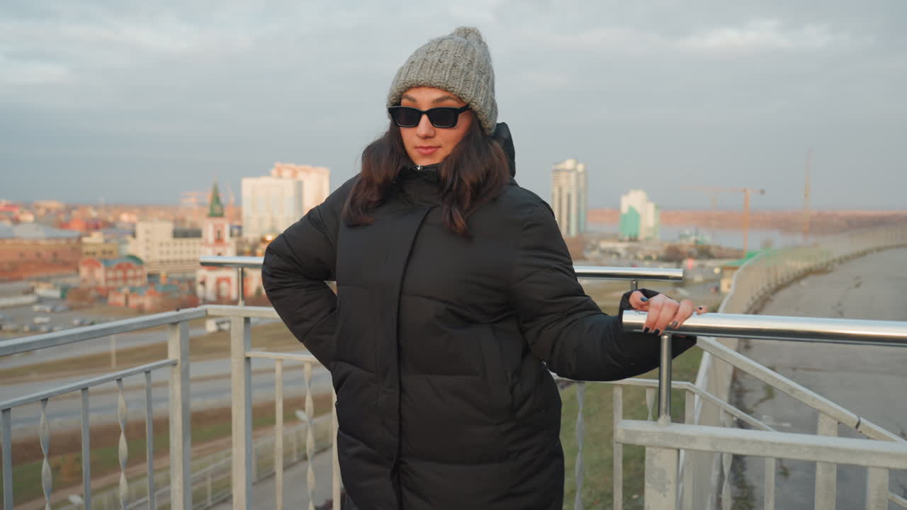 Donna che balla su una ringhiera di un belvedere cittadino, gesti delle braccia giocosi e posa fiduciosa con piumino nero e berretto di lana grigio, occhiali da sole abbassati, skyline dell'ora d'oro sullo sfondo, atmosfera esterna fresca.