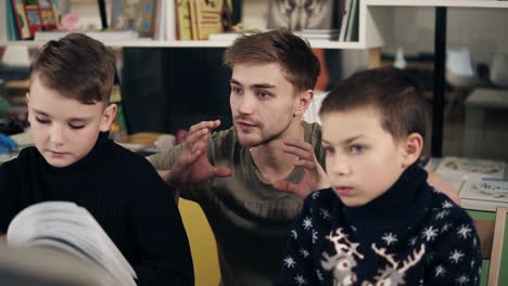 attractive young male teacher with couple tattooes on his arms explaining something to two little boys in warm cozy sweaters.