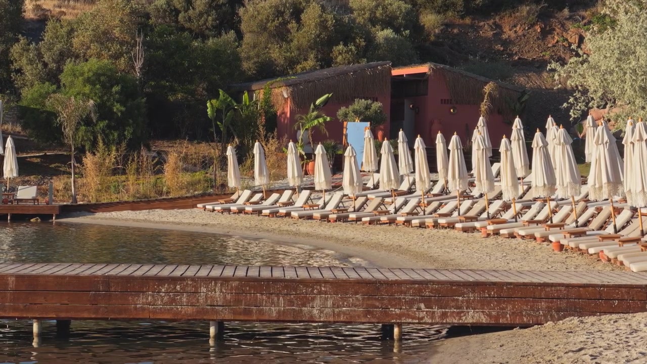 Una spiaggia deserta mostra file di lettini e ombrelloni accanto a un molo di legno e acqua calma.