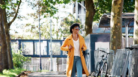 Fashionable-young-black-woman-wearing-a-hat,-sunglasses,-blue-jeans-and-a-yellow-pea-coat-walking-along-a-treelined-street-towards-camera,-listening-to-music-with-earphones-and-looking-around,-backlit,-low-angle