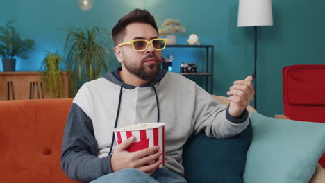 young man sits on sofa eating popcorn and watching interesting tv serial sport game online at home