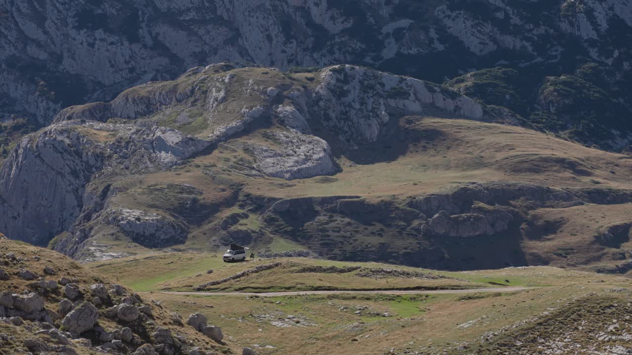 Static wide shot of rocky highland terrain with an overlanding vehicle parked below steep limestone cliffs in Durmitor National Park