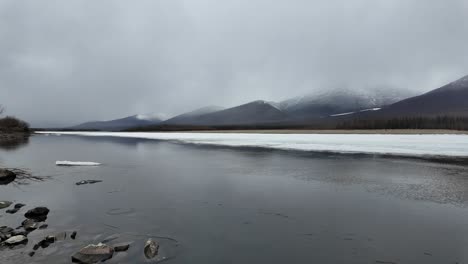 Langsame-Strömung-Einer-Weiteren-Hälfte-Des-Gefrorenen-Flusses-Vor-Dem-Hintergrund-Eines-Grauen-Himmels-Und-Schneebedeckter-Berge