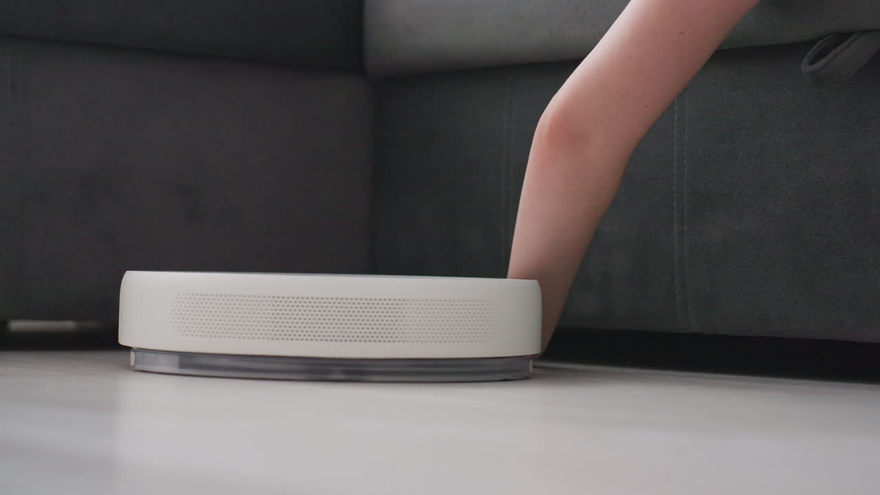 Robot vacuum cleaner approaches human hand resting on floor near couch then turns back and moves away, highlighting obstacle detection, smart home cleaning technology