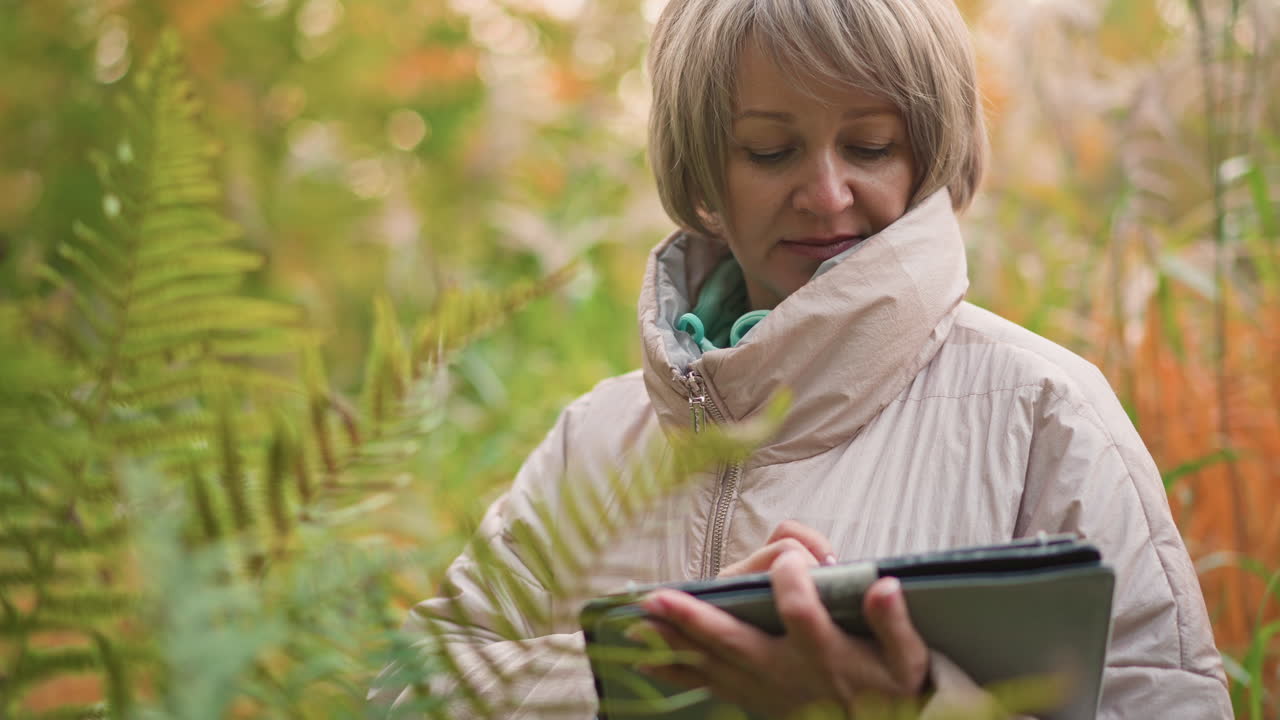 female scientist closely examining fern leaf while recording plant data during outdoor research in autumn forest, wearing warm jacket, surrounded by lush vegetation