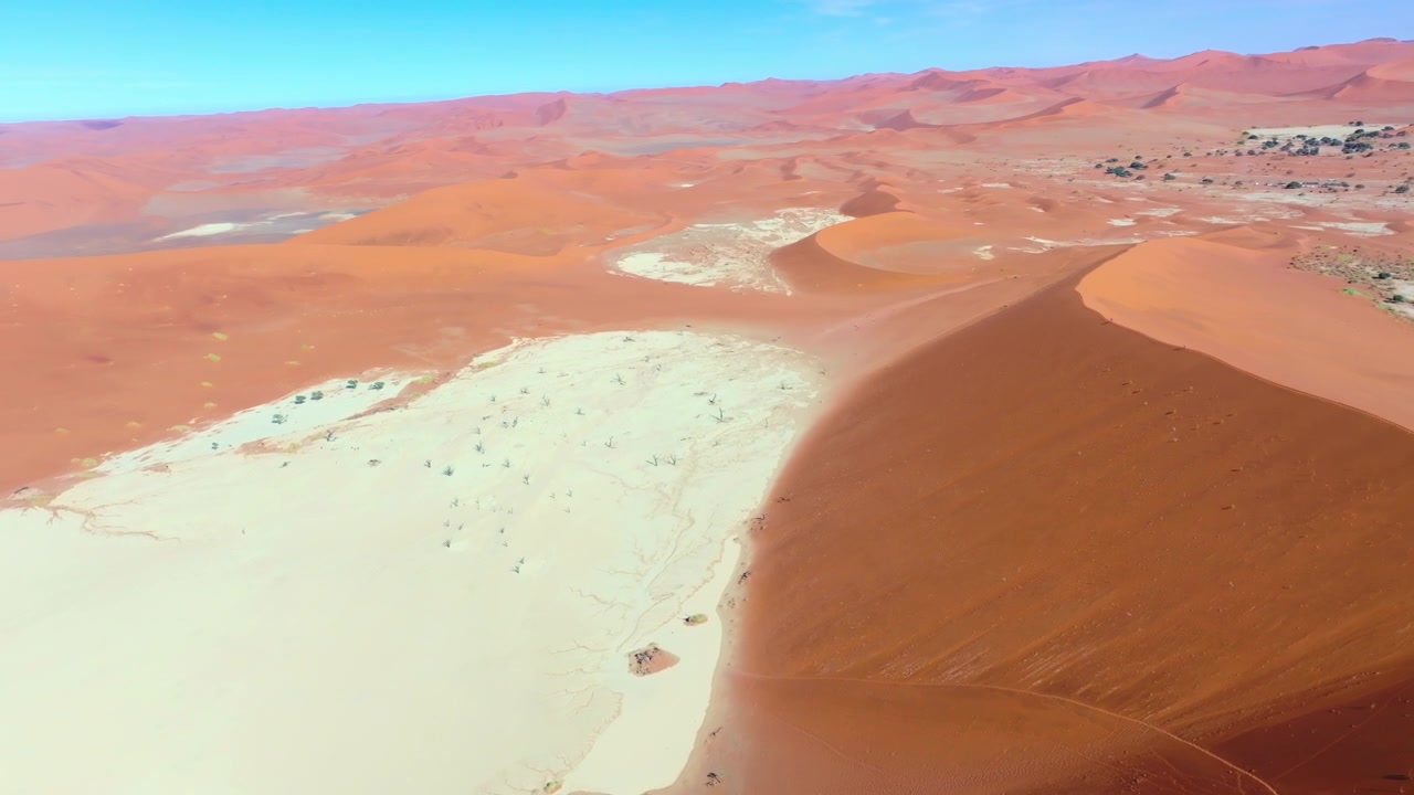 Vista aérea de turistas minúsculos caminhando pela planície branca de Deadvlei, ofuscados pelas dunas vermelhas de Sossusvlei. Pequenos visitantes são minimizados pelas imensas dunas vermelhas e pela planície branca pontilhada de árvores mortas.