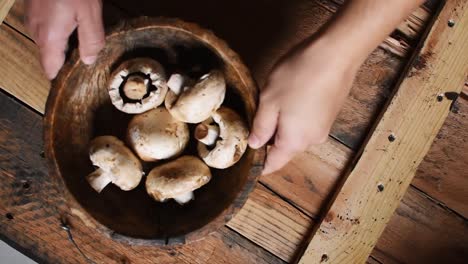 man's-hands-putting-a-wooden-bowl-with-white-mushrooms-inside-on-rustic-wooden-texture-as-background,high-angle,-steady-cam