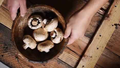 man's hands putting a wooden bowl with white mushrooms inside on rustic wooden texture as background,high angle, steady cam