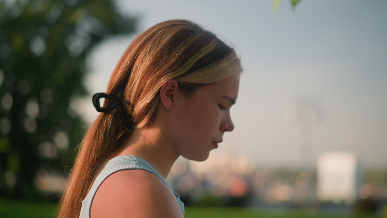 Side view of young lady looking pensive while sunlight reflects on her face, with graceful movement like skating, blurred natural background includes greenery, trees, and subtle cityscape in distance