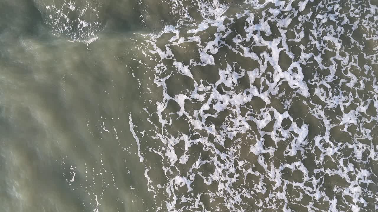 Top-down drone footage captures gentle ocean waves rolling onto a sandy beach in Port Douglas, Australia, under soft natural daylight with smooth camera movement