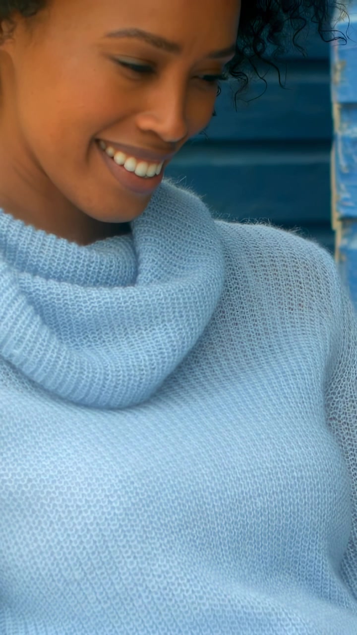 Vídeo vertical: Mulher sorrindo inclinando a cabeça após uma deixa fora da tela, na persiana, usando um suéter gola alta azul claro.
